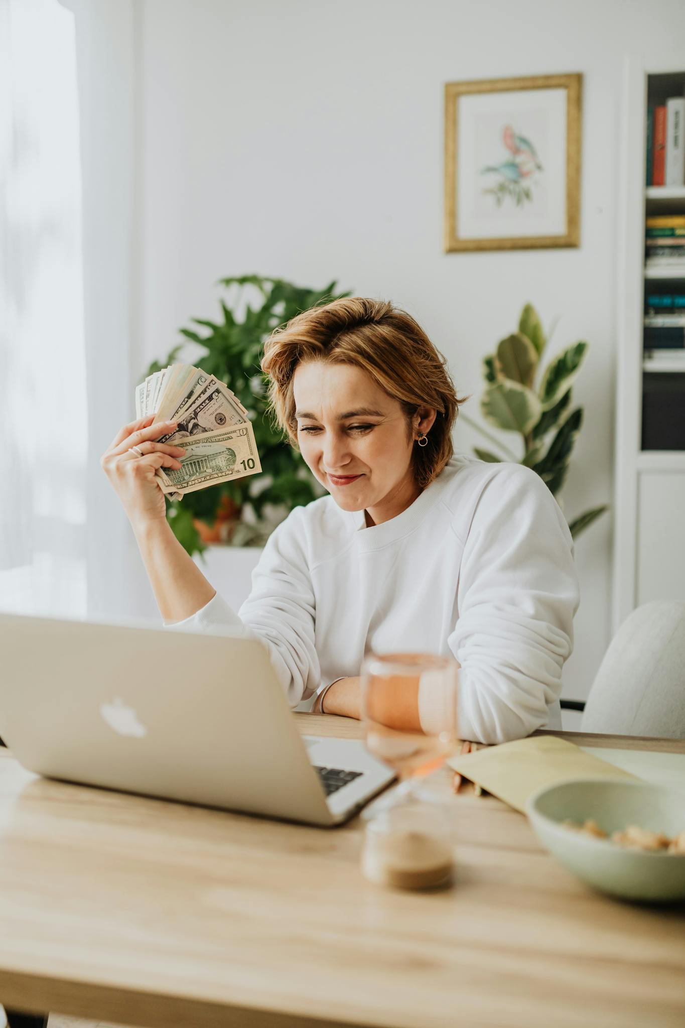 Woman seated at a desk holding dollar bills, working on a laptop, showcasing financial success from home.